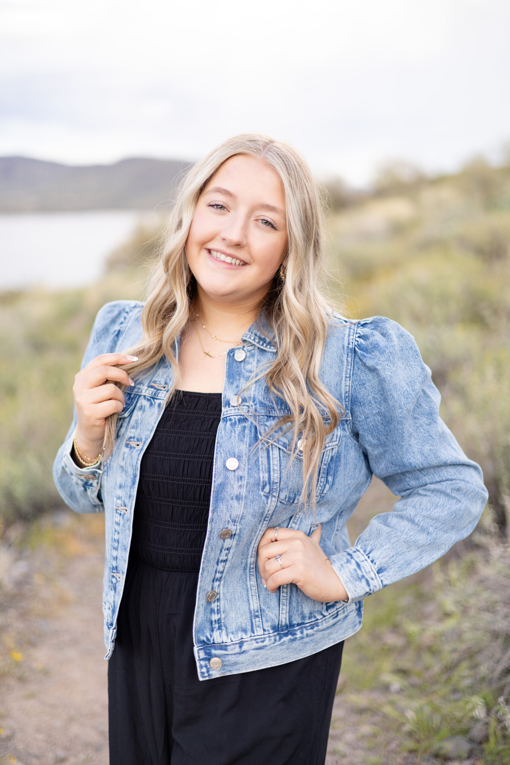 Senior in denim jacket and black jumper smiles at Lake Pleasant for senior session in Peoria, AZ.