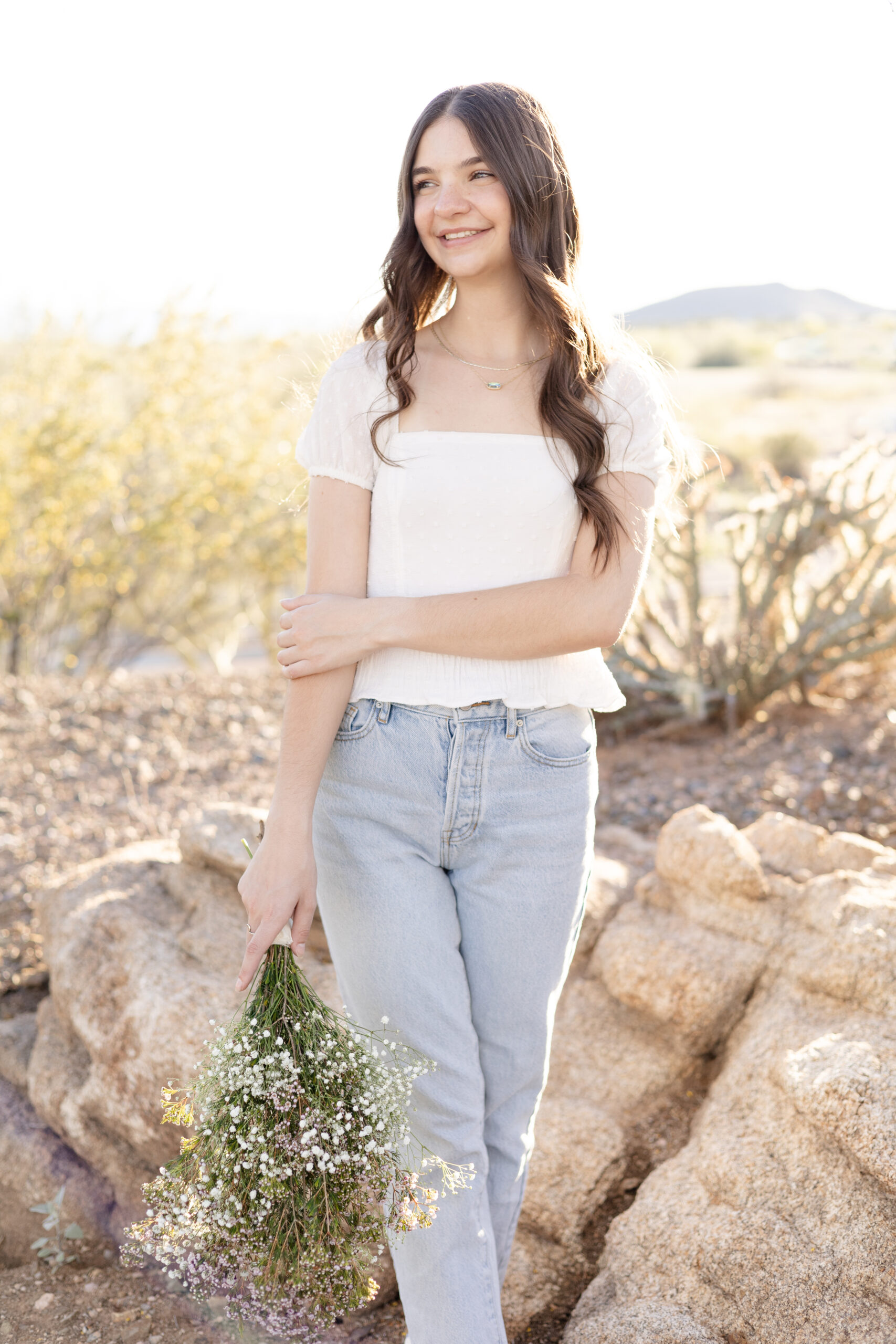 Senior stands with bouquet of flowers in denim jeans and white top at senior session in Vistancia.