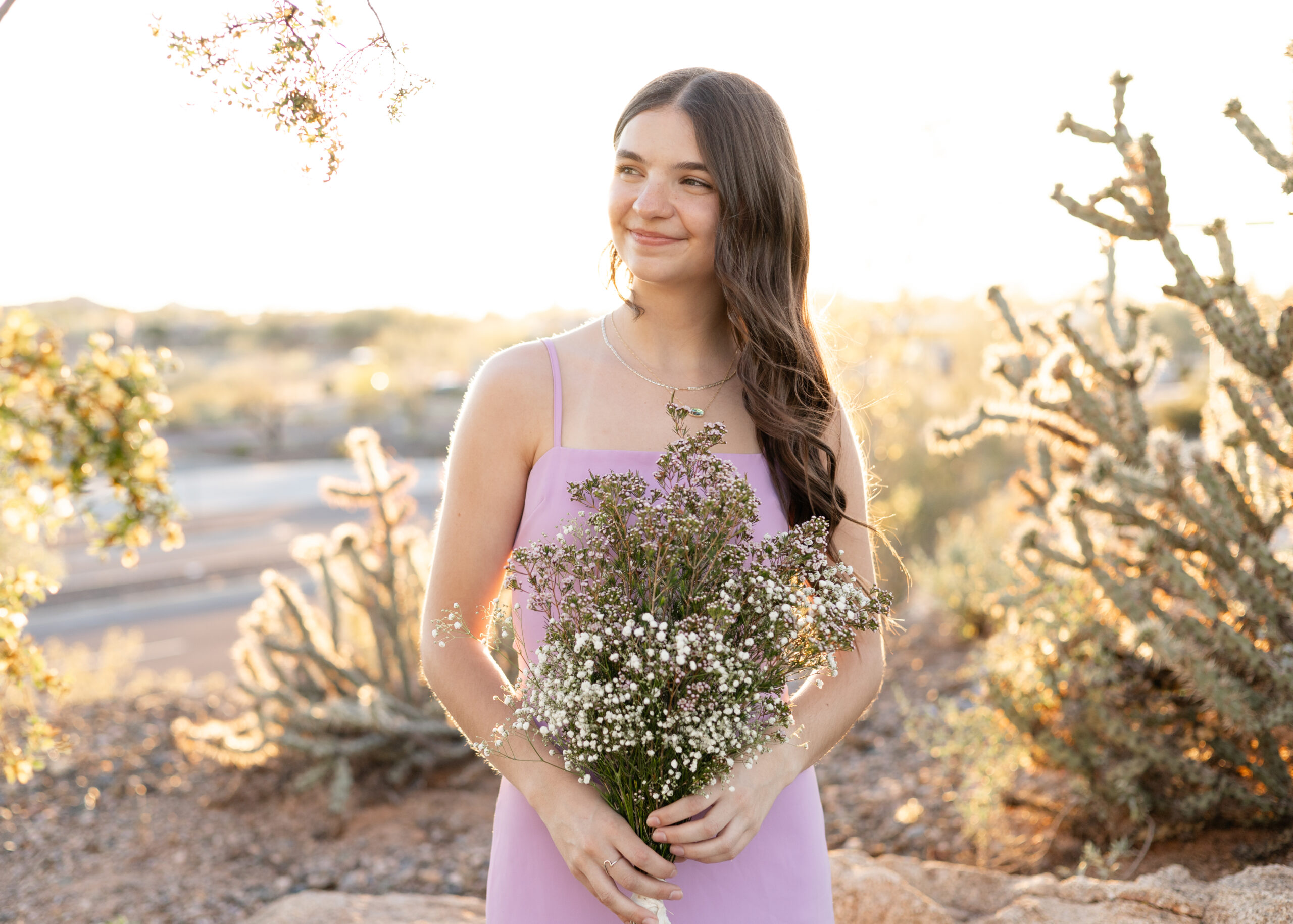 Senior girl in lavender dress holds bouquet of flowers in desert at sunset.