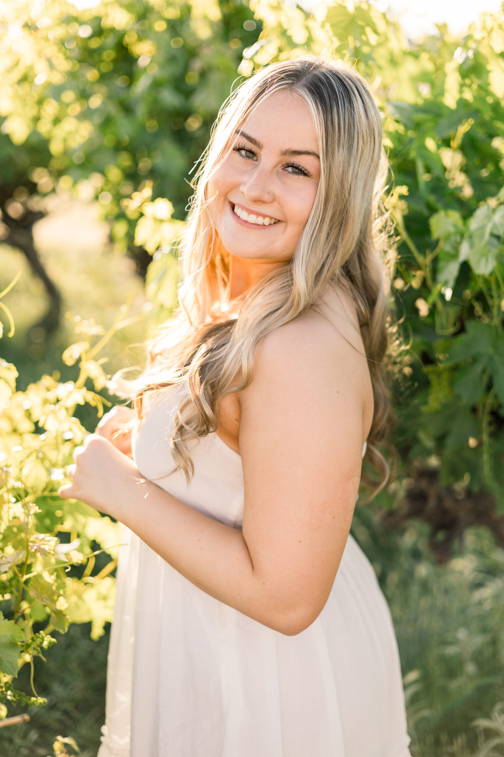 Senior in white dress stands among vineyard rows in Glendale, AZ.